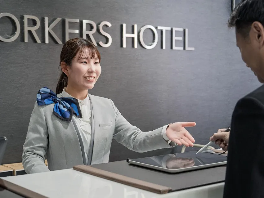 「WORKERS HOTEL」のロゴを背景に、笑顔で接客を行うホテルスタッフ。