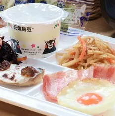 焼き魚、ベーコンエッグ、きんぴらごぼう、そして熊本のご当地キャラクター「くまモン」が描かれた納豆がプレートに並び、一日の活力を与えてくれる充実した朝食。