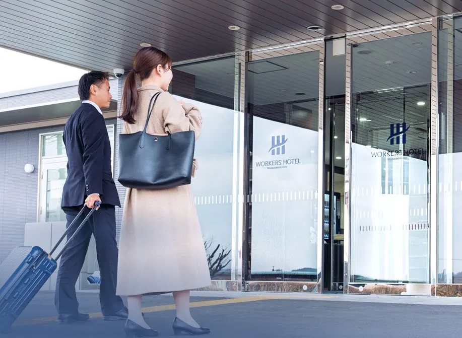 スーツケースを引く男性とトートバッグを肩にかけた女性が、ガラス張りの「WORKERS HOTEL」の入り口へ向かって歩いている様子。